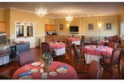 Dining room with round tables set for meals, pink tablecloths, chandeliers, and a buffet/service area.