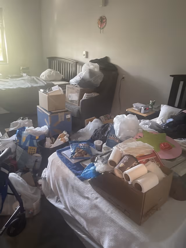 A cluttered bedroom with a bed covered in various items including paper towels, bags, snacks, and other miscellaneous objects. There is a chair and a small table with an alarm clock showing 2:35. The room has a window letting in natural light and a wall clock hanging above the chair.