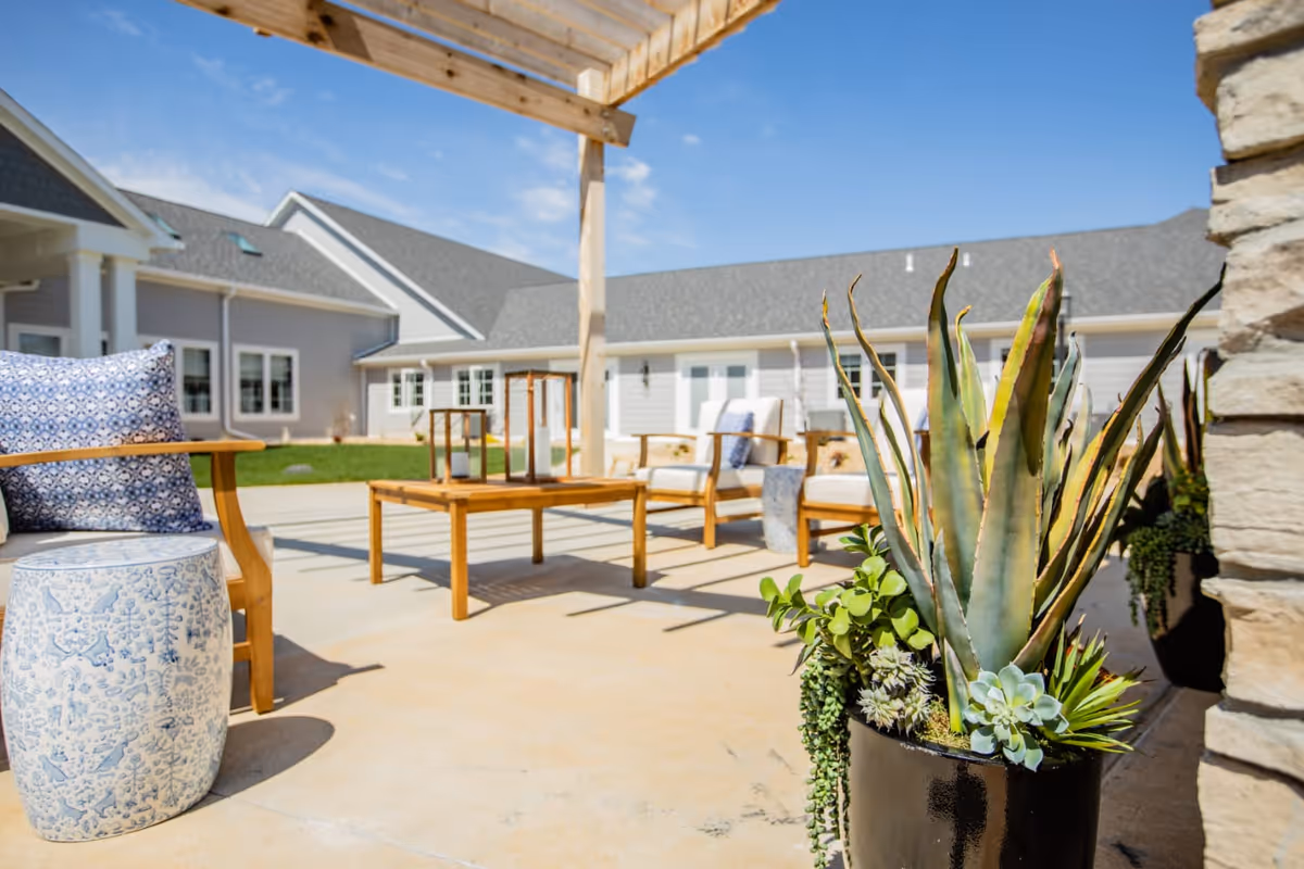 Outdoor patio area at Cedarhurst Senior Living of West Plains featuring wooden chairs with cushions, a wooden coffee table with lanterns, a blue and white ceramic stool, and a large black planter with various succulents under a wooden pergola. The background shows the exterior of the senior living facility building under a clear blue sky.