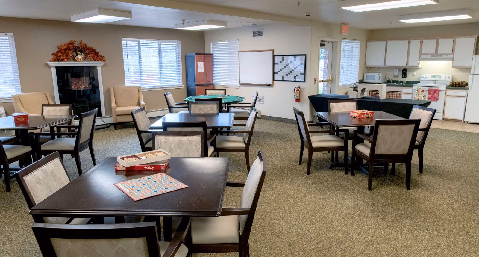 A spacious common room with several tables and chairs arranged for group activities. One table has a Scrabble game set up. There are two beige armchairs near a decorative fireplace with autumn-themed decor on the mantel. In the background, there is a kitchen area with white cabinets, a stove, microwave, and countertops. The room has multiple windows with blinds and a whiteboard and crossword puzzle board on the wall.