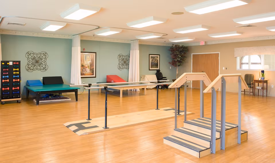 A spacious rehabilitation room with wooden flooring, parallel bars for walking exercises, a set of stairs with handrails, exercise mats, a rack of colorful dumbbells, and a chair near a window with a small table and plant.