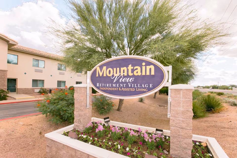 Outdoor view of the entrance sign for Mountain View Retirement Village, an independent and assisted living facility, with a building and desert landscaping in the background under a partly cloudy sky.