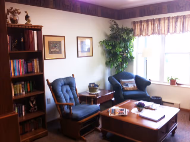 A cozy living room with two blue cushioned armchairs, a wooden coffee table with magazines and a small plant, a wooden bookshelf filled with books, framed artwork on the wall, a tall green plant in the corner, and a window with curtains letting in natural light.