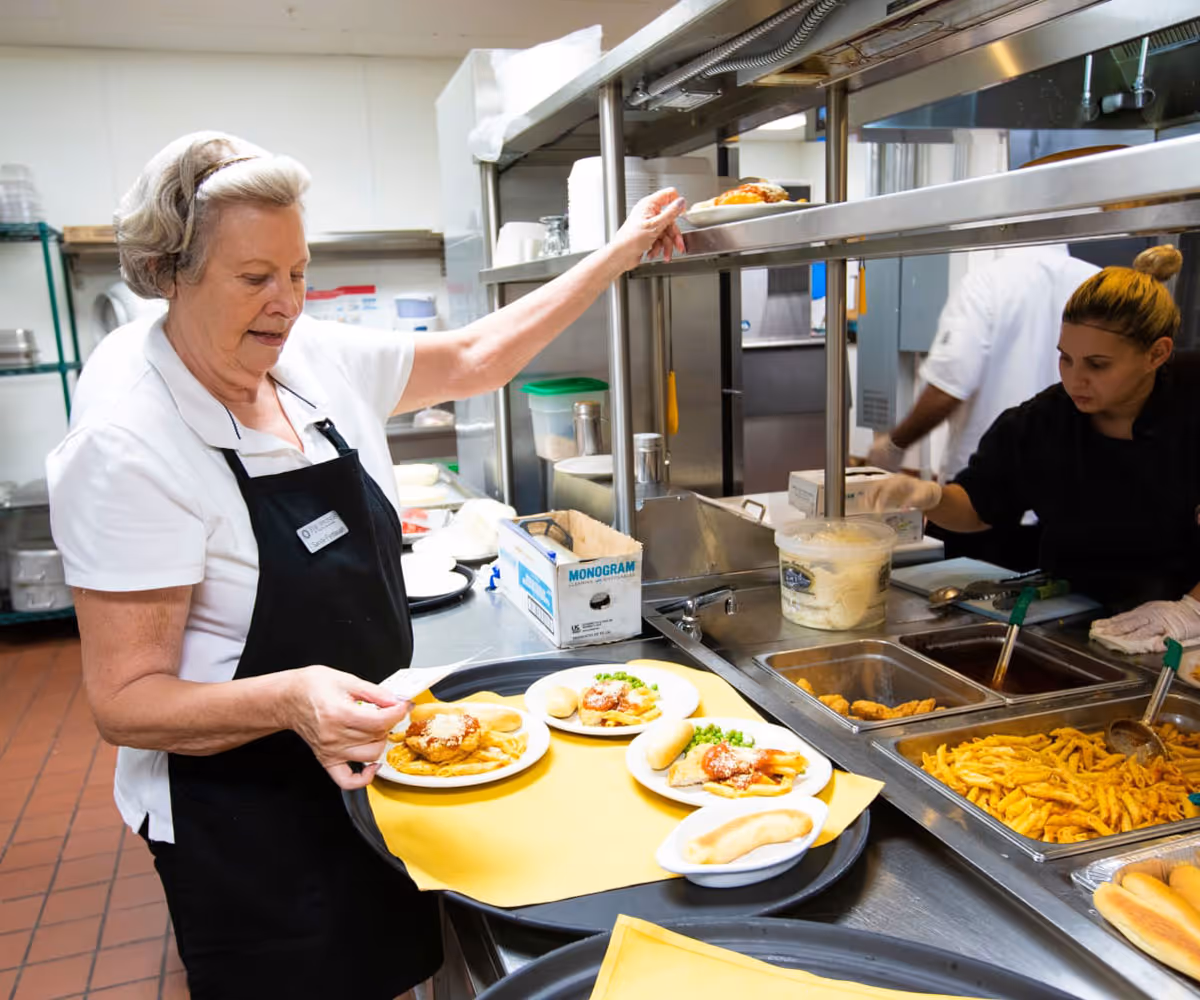 A woman wearing a white shirt and black apron serves plates of food including pasta, bread rolls, and peas in a kitchen setting. Another person in black attire is seen working behind the serving counter with trays of food such as pasta and sauce.