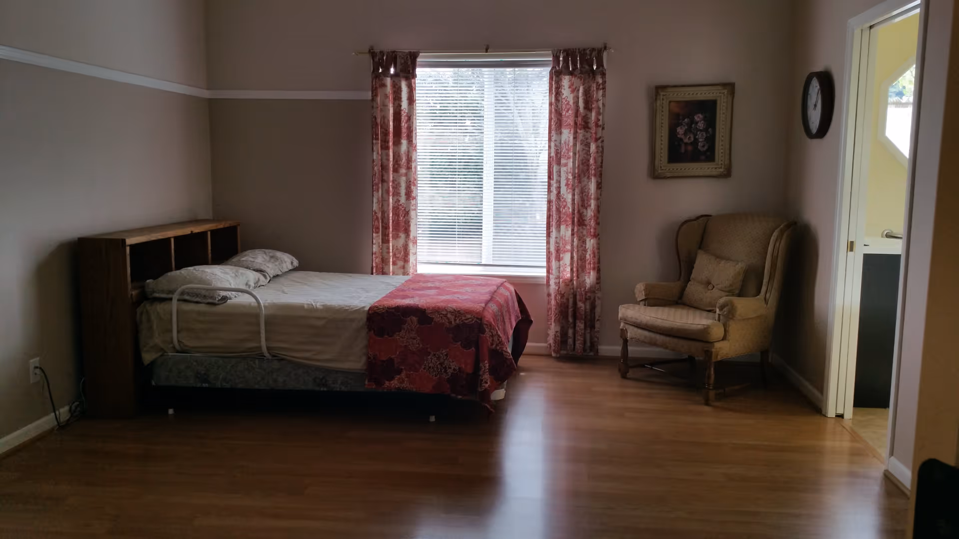 A bedroom with a single bed against the left wall, featuring a wooden headboard with shelves. The bed is made with white sheets, a pillow, and a red patterned blanket. A large window with floral curtains is centered on the back wall. To the right of the window is a beige upholstered armchair with a cushion. A framed floral painting hangs on the wall above the chair. A clock is mounted on the right wall near an open doorway leading to another room. The floor is wooden.