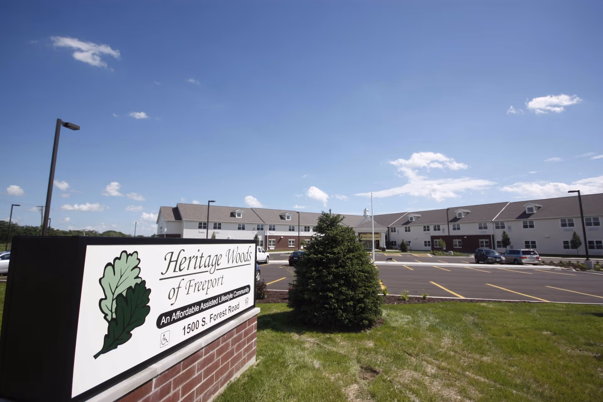 Exterior view of Heritage Woods of Freeport, an assisted living community, showing a large building with multiple windows and a parking lot in front under a blue sky with scattered clouds. A sign in the foreground displays the facility's name and address.