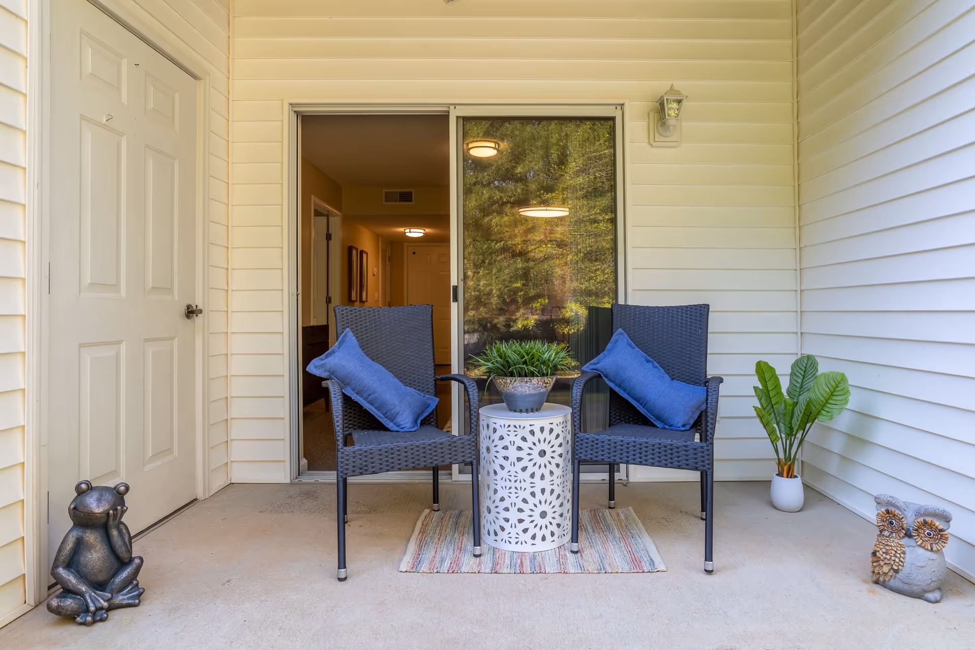 A cozy outdoor patio area with two black wicker chairs each with a blue cushion, a small white decorative round table with a potted plant on top, a striped rug underneath, and two small statues of a frog and two owls on the concrete floor. There is a sliding glass door leading inside and a white door to the left. The walls are covered with light-colored siding.