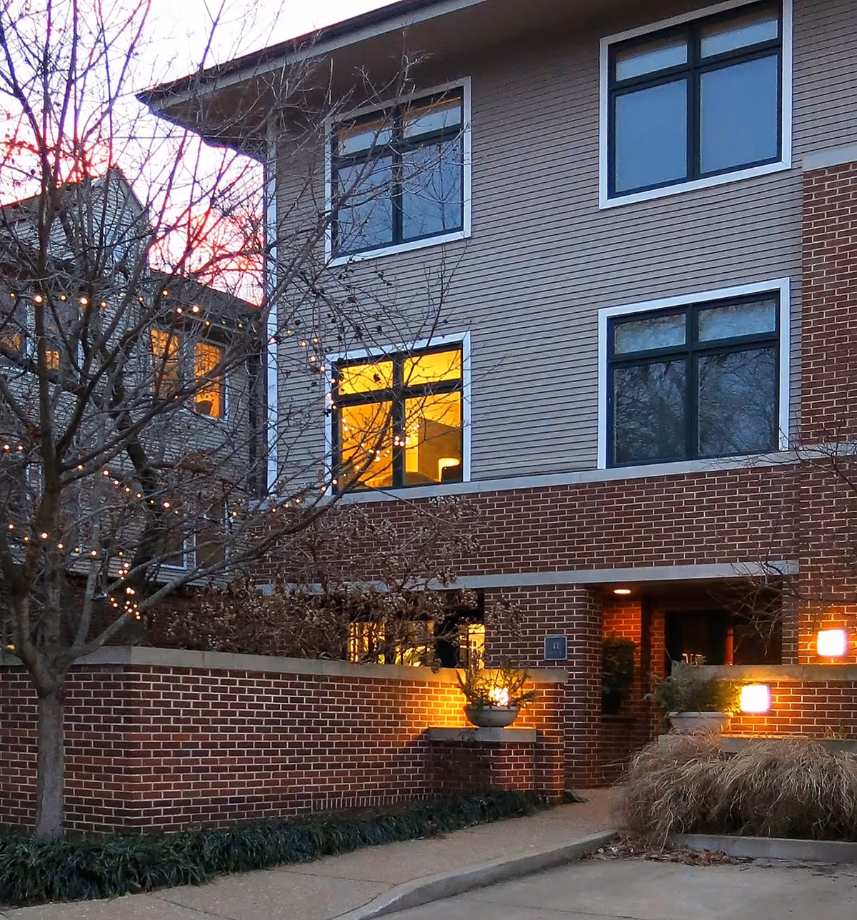 Exterior view of a senior living facility building at dusk with lit windows, brick and siding facade, bare trees with string lights, and a walkway leading to the entrance.