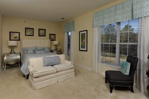 Sunlit bedroom with an upholstered bed, a small loveseat at the foot, a patterned chair by a large window, and bedside tables with lamps.