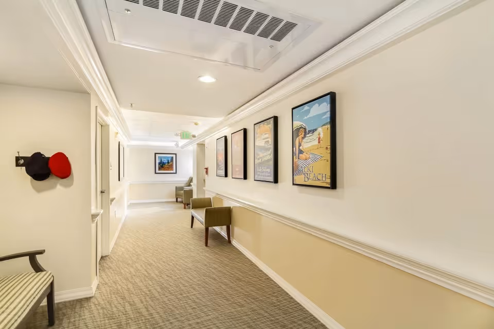 A well-lit hallway in a senior living facility with beige walls and carpeted floor. The right wall features four framed pictures, including one of a beach scene. There are two green cushioned benches along the right wall and a coat rack with two hats on the left wall. At the end of the hallway, there are two armchairs and a framed picture on the wall.