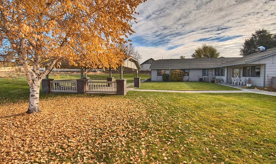 Single-story residential care building with a grassy courtyard, a gazebo, and a tree shedding autumn leaves.