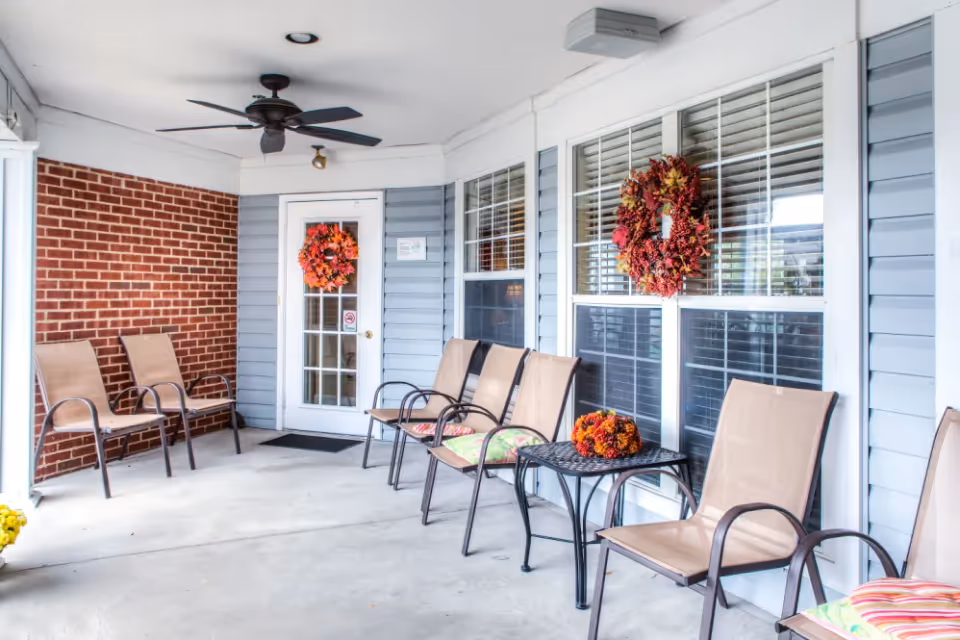 Covered outdoor porch area with several beige mesh chairs arranged along the walls, a small black metal table with a fall-themed centerpiece, two fall wreaths hanging on the windows and door, a ceiling fan, and a brick and light blue siding exterior.