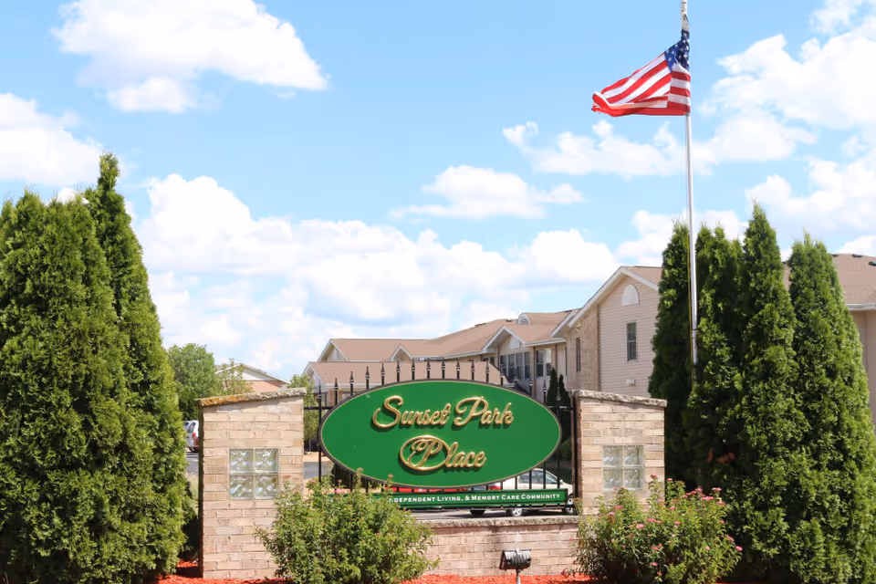 Entrance sign for Sunset Park Place, an independent living and memory care community, surrounded by greenery and tall trees with a clear blue sky and an American flag on a flagpole in the background.