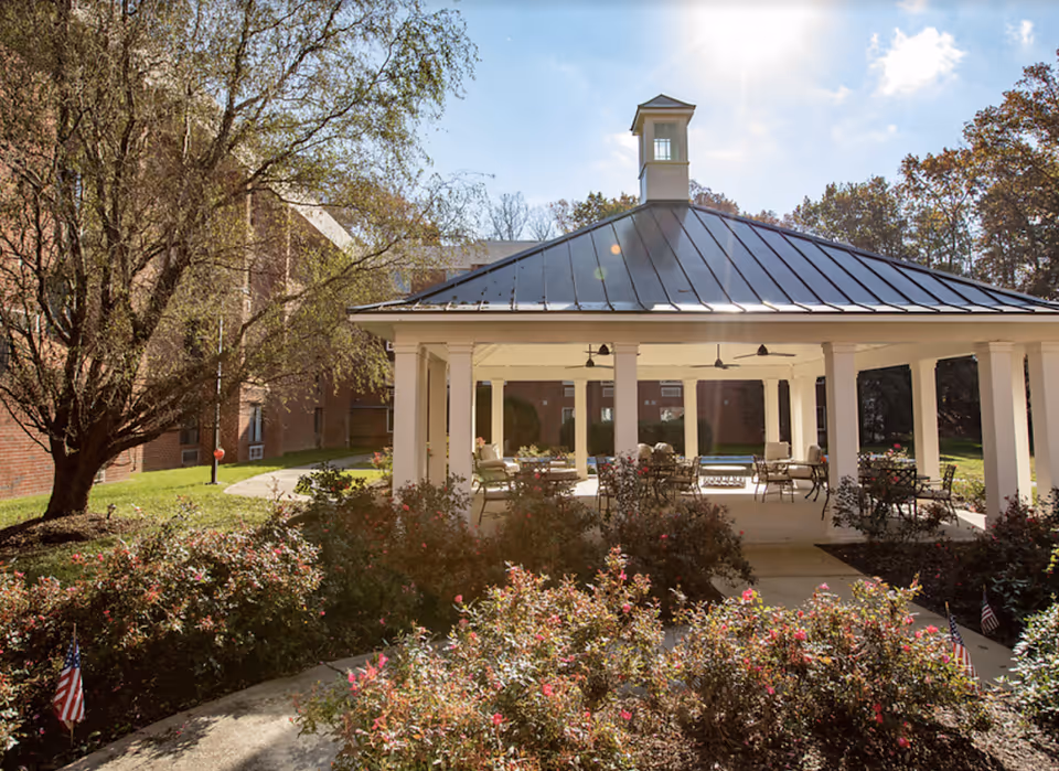 Covered outdoor pavilion with seating in a landscaped courtyard surrounded by trees and flowering shrubs.