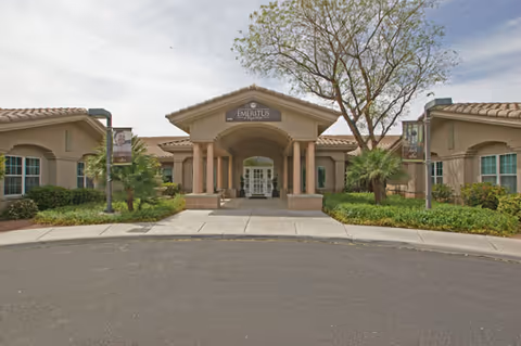 Front exterior view of a single-story senior living facility building with a covered entrance supported by columns, surrounded by landscaped greenery and trees under a partly cloudy sky.