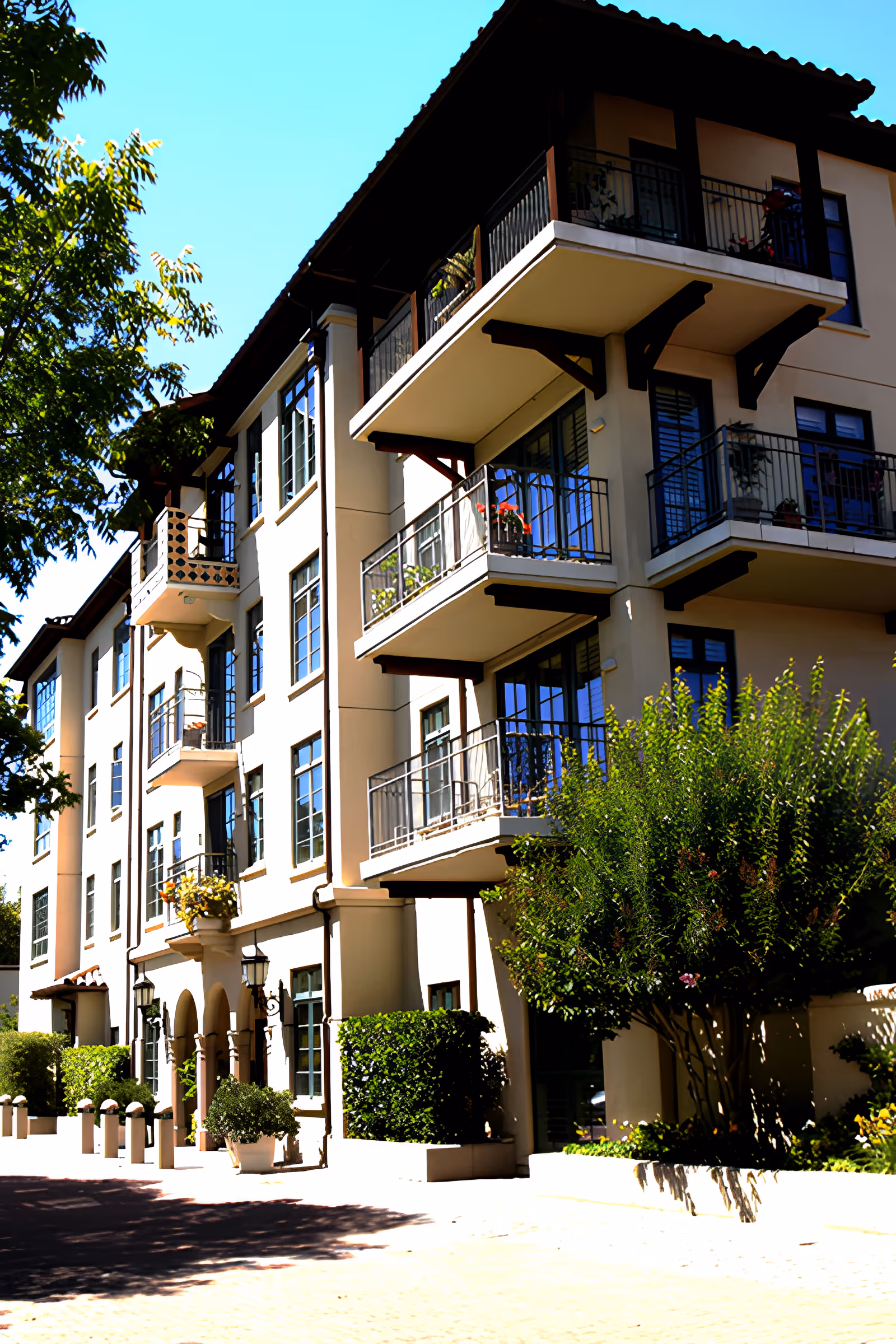 Sunlit multi-story residential building facade with balconies, arched entrance, and landscaped shrubs.
