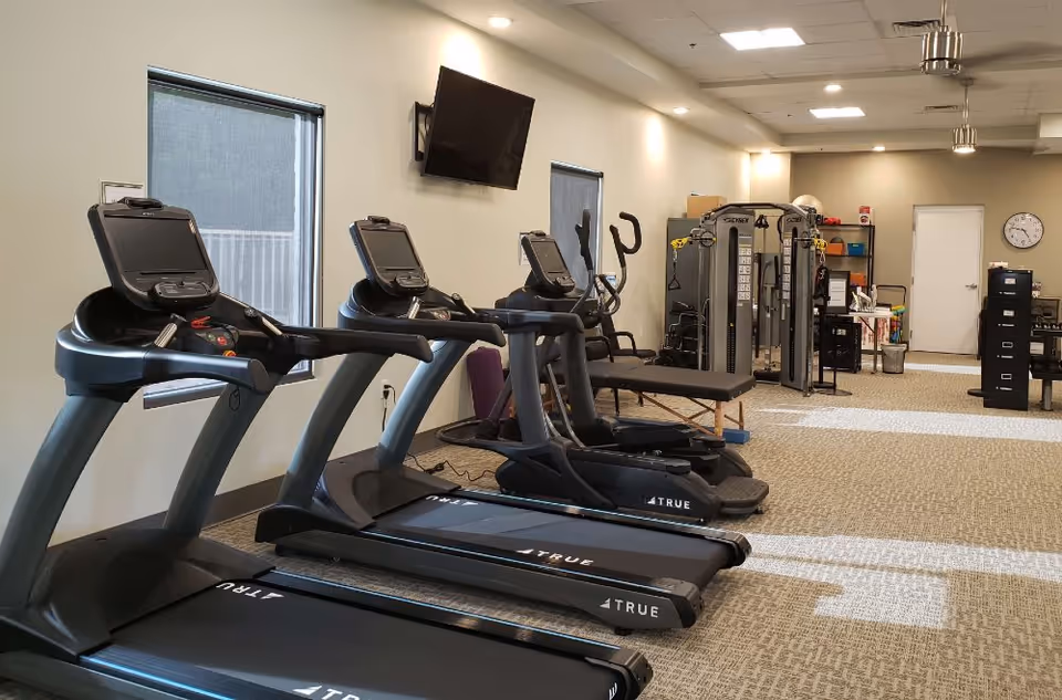 Interior view of a fitness room with three treadmills, an elliptical machine, a multi-functional exercise machine, a wall-mounted TV, and windows with blinds. The room has carpeted flooring and bright overhead lighting.