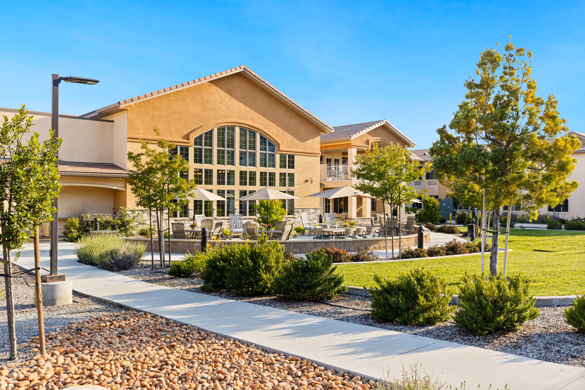 Outdoor view of Ivy Park At Murrieta facility showing a well-maintained garden area with green grass, trees, shrubs, and a paved walkway. The building has large arched windows and a patio area with tables, chairs, and umbrellas under a clear blue sky.