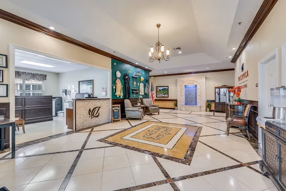 Spacious and well-lit senior living facility lobby with a decorative tiled floor, a chandelier hanging from the ceiling, a water feature on the far wall, two armchairs with a small table, an American flag, and a reception desk with a leaf logo. The walls are adorned with various decorations and plaques, and there is a water dispenser on the right side.