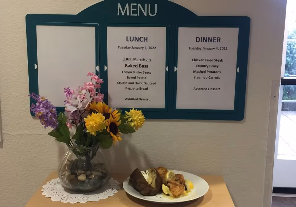 Wall-mounted daily menu above a small table holding a vase of flowers and a plate of food.