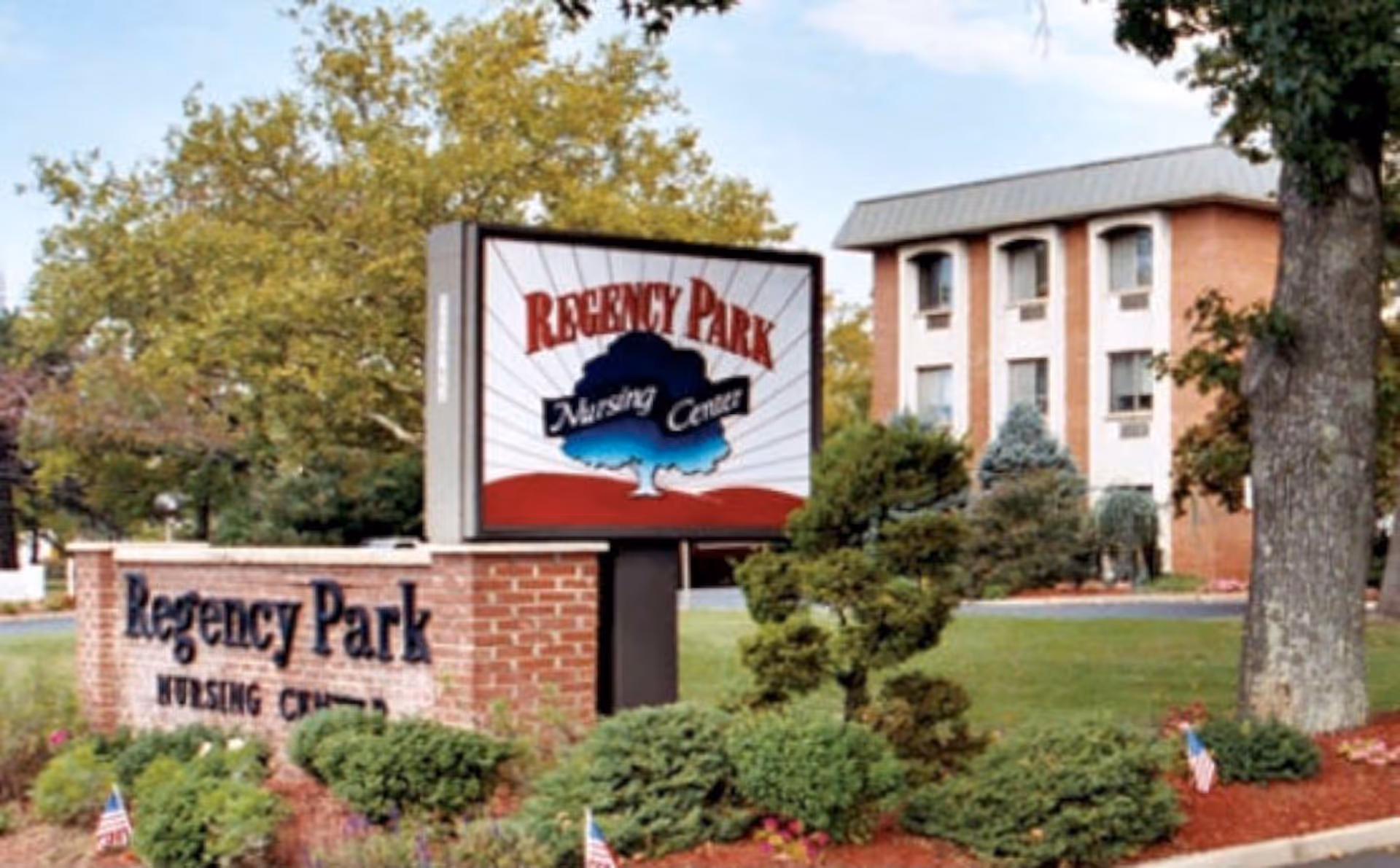 Brick entrance sign and illuminated marquee reading 'Regency Park Nursing Center' in front of a three-story brick building surrounded by trees and landscaping.