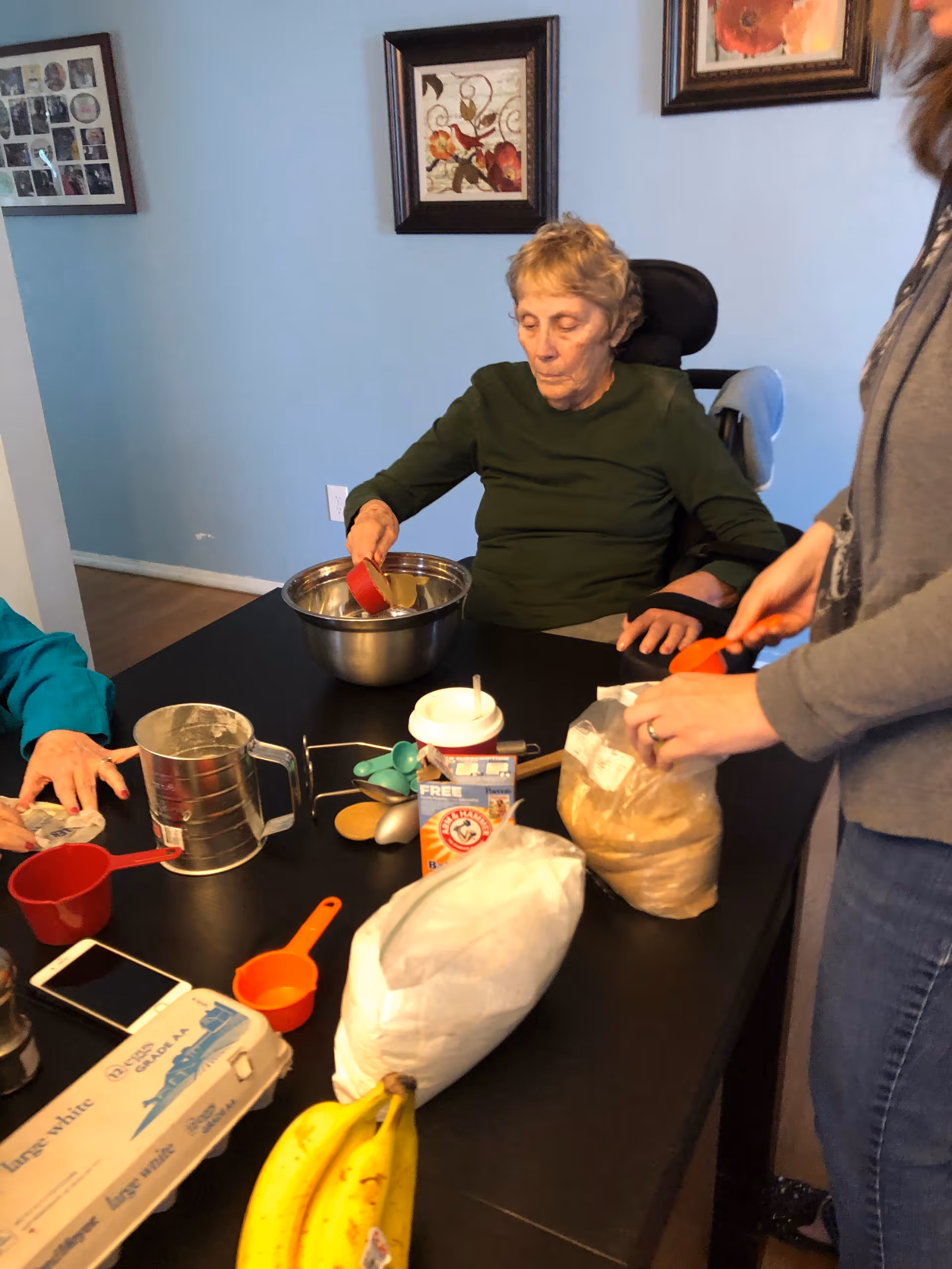 An elderly woman seated at a table mixes ingredients in a bowl while another person measures brown sugar and baking supplies sit on the table.