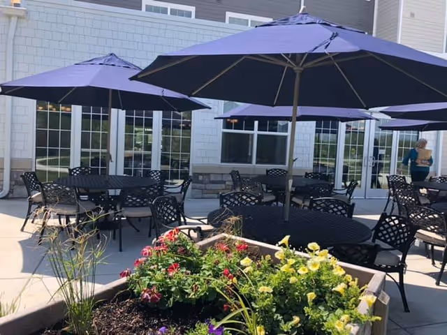 Outdoor patio area with multiple black metal tables and chairs, each shaded by large purple umbrellas. In the foreground, there is a raised garden bed with colorful flowers. The background shows a building with large windows and a person walking near the entrance.