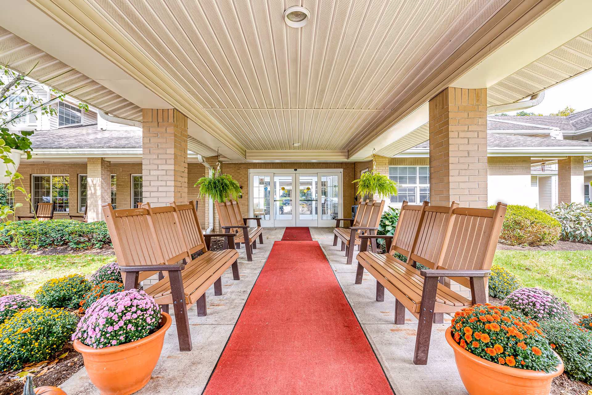 Covered entrance area of a senior living facility with a red carpet runner down the center, flanked by wooden benches on both sides. There are hanging green plants and large pots with colorful flowers near the benches. The building has beige brick pillars and white double doors at the end of the walkway.