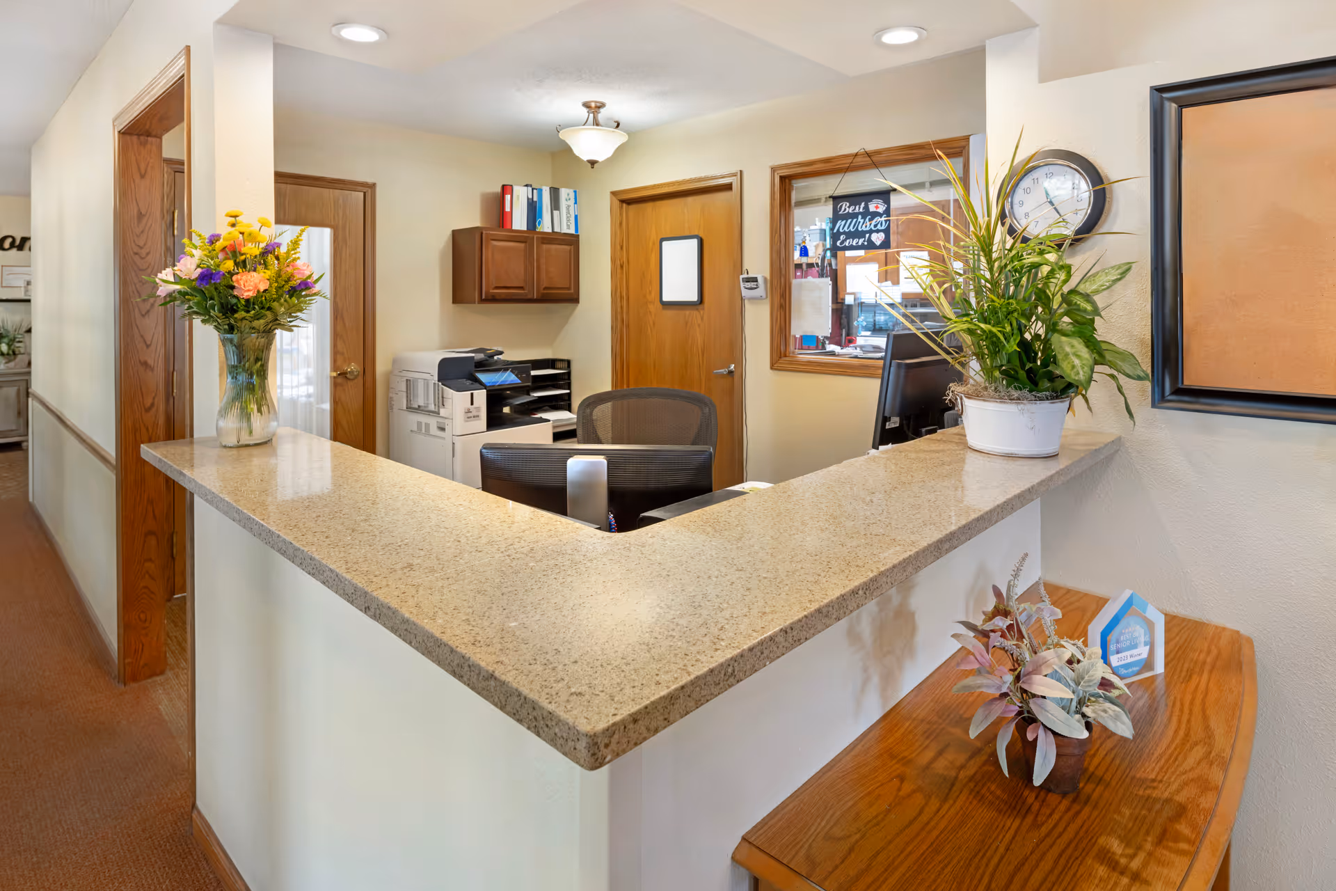 Reception area with a beige countertop desk, two flower arrangements, a clock on the wall, a window with a sign that reads 'Best nurses ever!', office chairs, a printer, and wooden doors in the background.