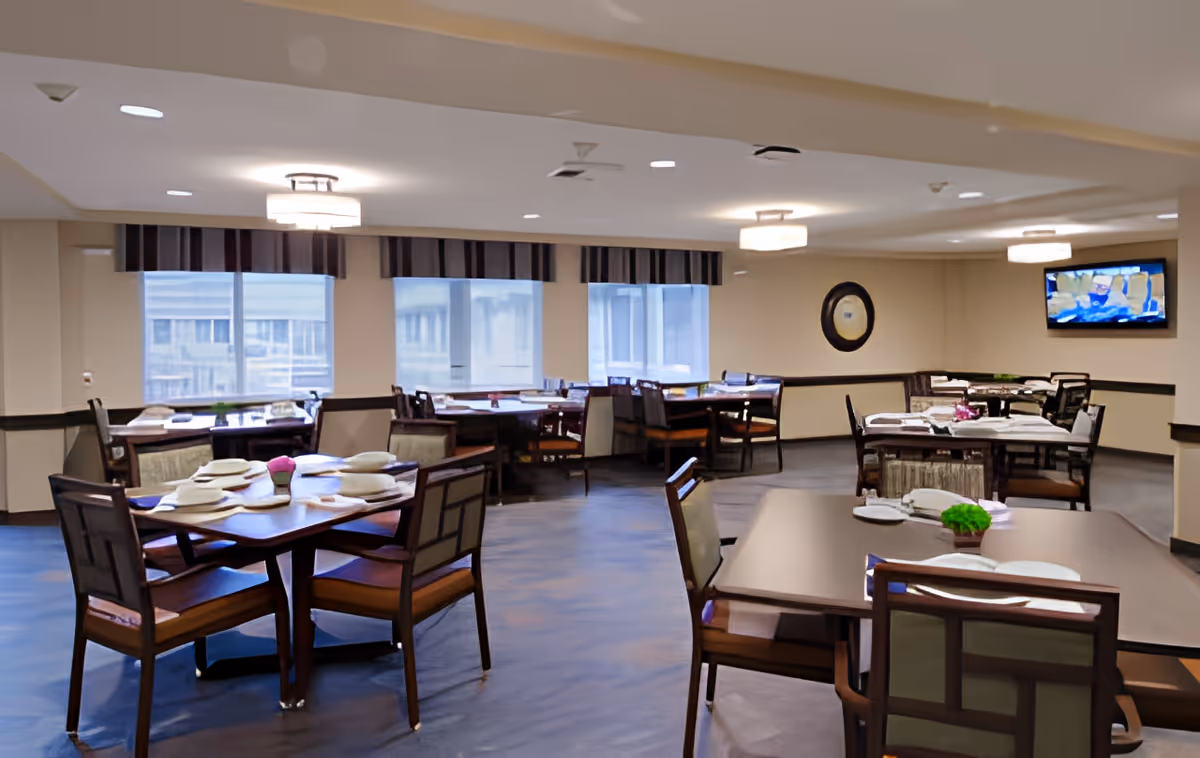 A dining room with multiple tables set with plates, napkins, and small decorative plants. The room has large windows with striped valances, ceiling lights, a wall-mounted TV, and a round mirror on the wall.