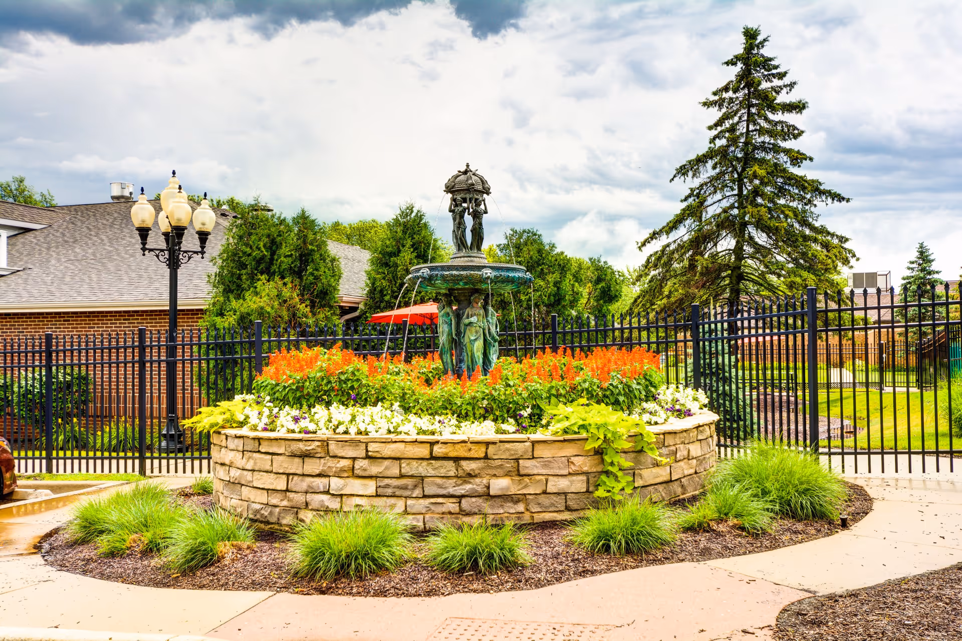 Outdoor garden area with a decorative stone fountain surrounded by colorful flowers and greenery, black metal fencing, a tall pine tree, and a brick building in the background under a partly cloudy sky.