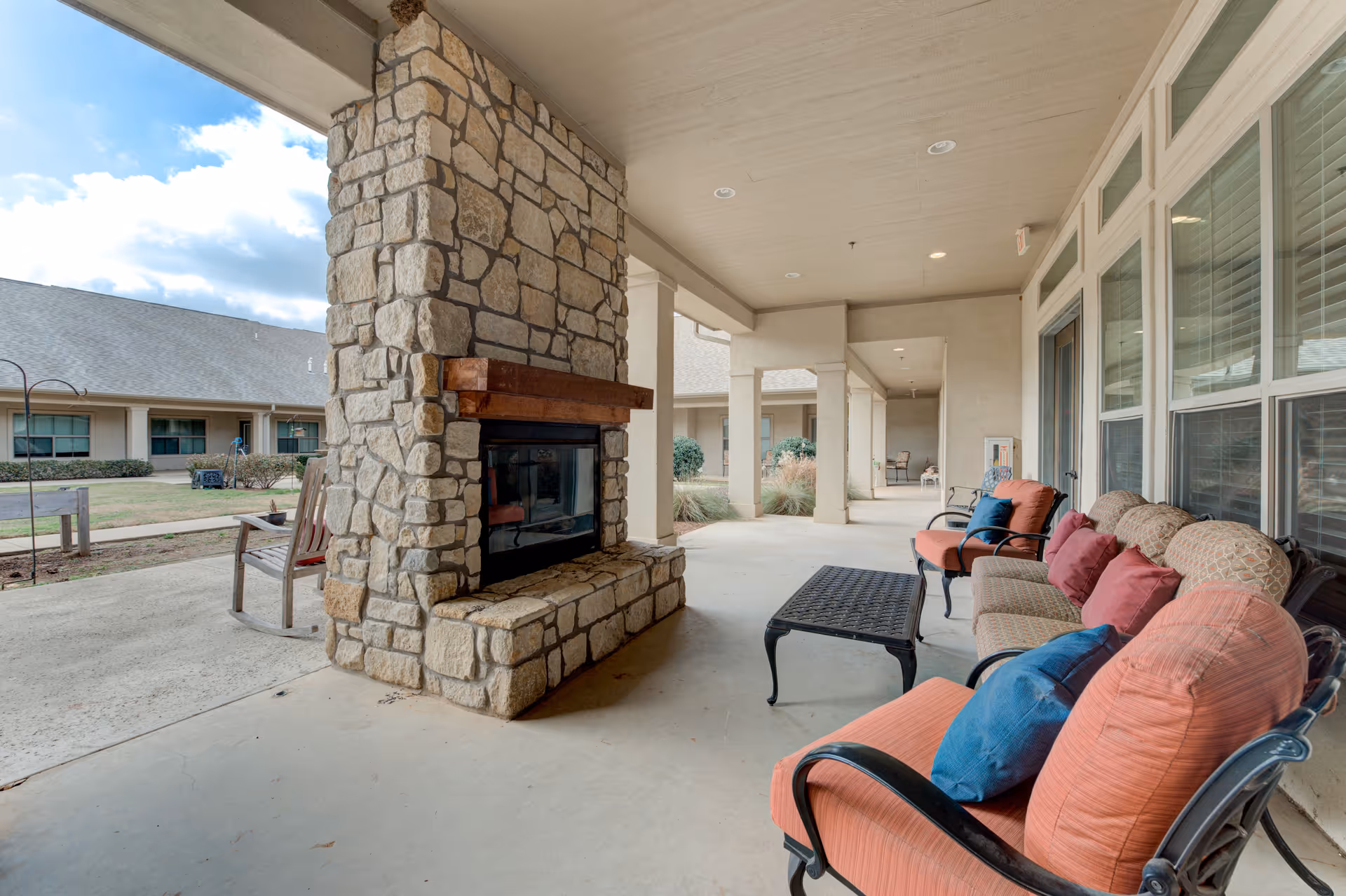 Covered outdoor patio area with cushioned seating including armchairs and a sofa, a stone fireplace with a wooden mantel, and a black metal coffee table. The patio overlooks a courtyard with grass and shrubs, and the building exterior with windows and doors is visible.