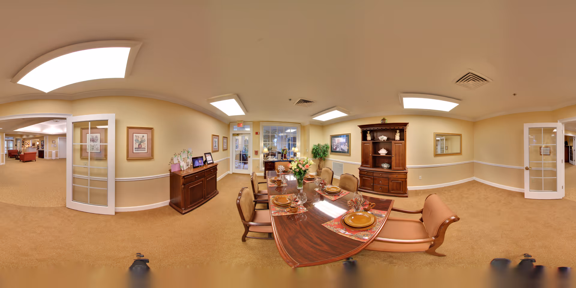 A well-lit dining room in a senior living facility with a long wooden dining table set with plates, glasses, and floral centerpieces. The room has beige walls, carpeted floor, and ceiling lights. There are wooden cabinets and framed artwork on the walls. Glass-paneled double doors open to a hallway and other rooms.