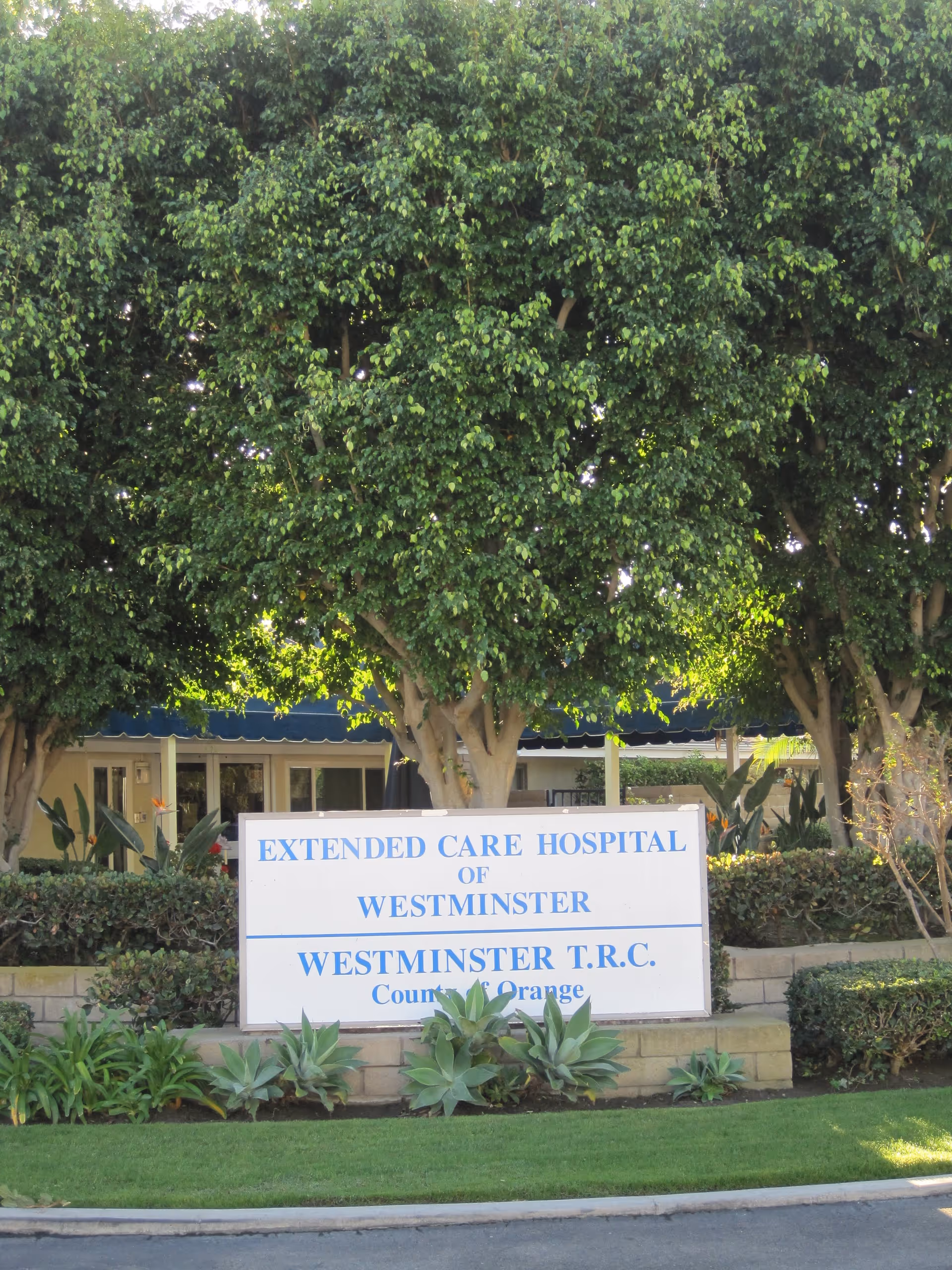 Outdoor view of the entrance area of Extended Care Hospital of Westminster with a large white sign displaying the facility's name surrounded by green plants and trees.