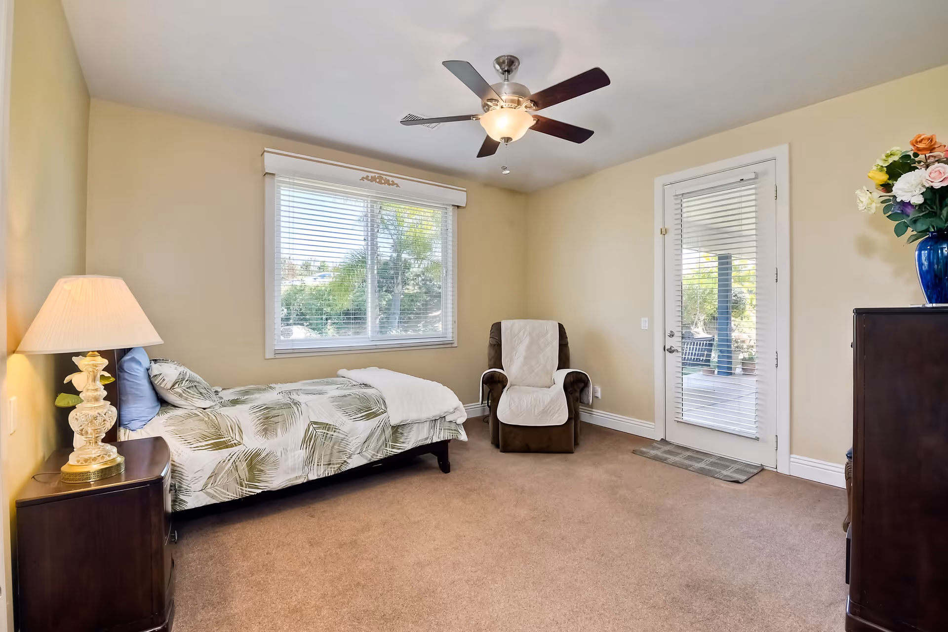 A tidy bedroom with a single bed, nightstand and lamp, a recliner, ceiling fan, window with blinds and a glass door to the outside.
