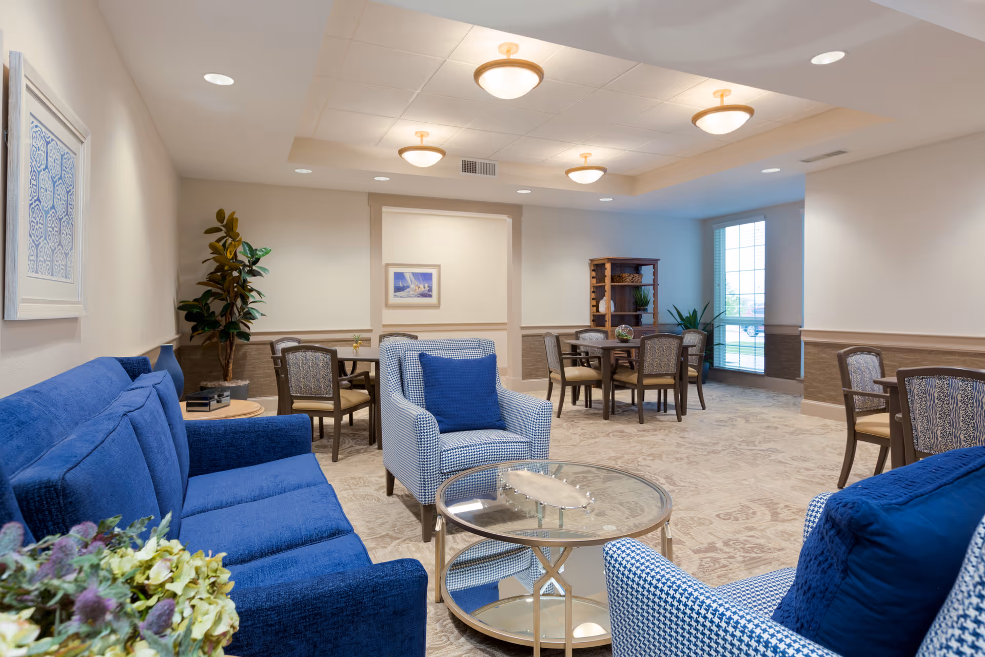A cozy senior living common area with blue upholstered sofas and armchairs, a glass coffee table, round wooden tables with chairs, potted plants, framed artwork on the walls, and large windows letting in natural light.