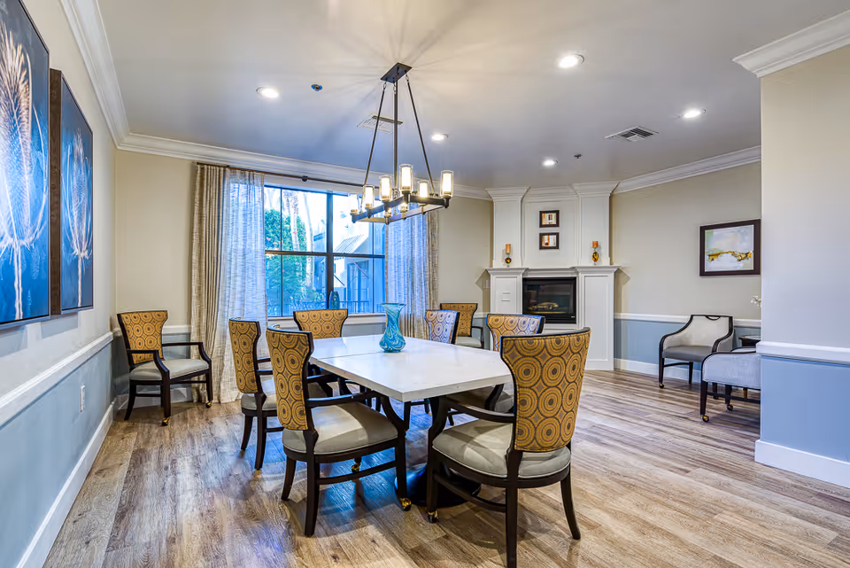 A well-lit dining room with a rectangular white table surrounded by eight patterned chairs. A modern chandelier hangs above the table. There is a large window with curtains letting in natural light, and a fireplace with decorative items on the mantel. The room has light-colored walls with white trim and wood flooring. Two armchairs and a small table are placed in a corner, and artwork is displayed on the walls.