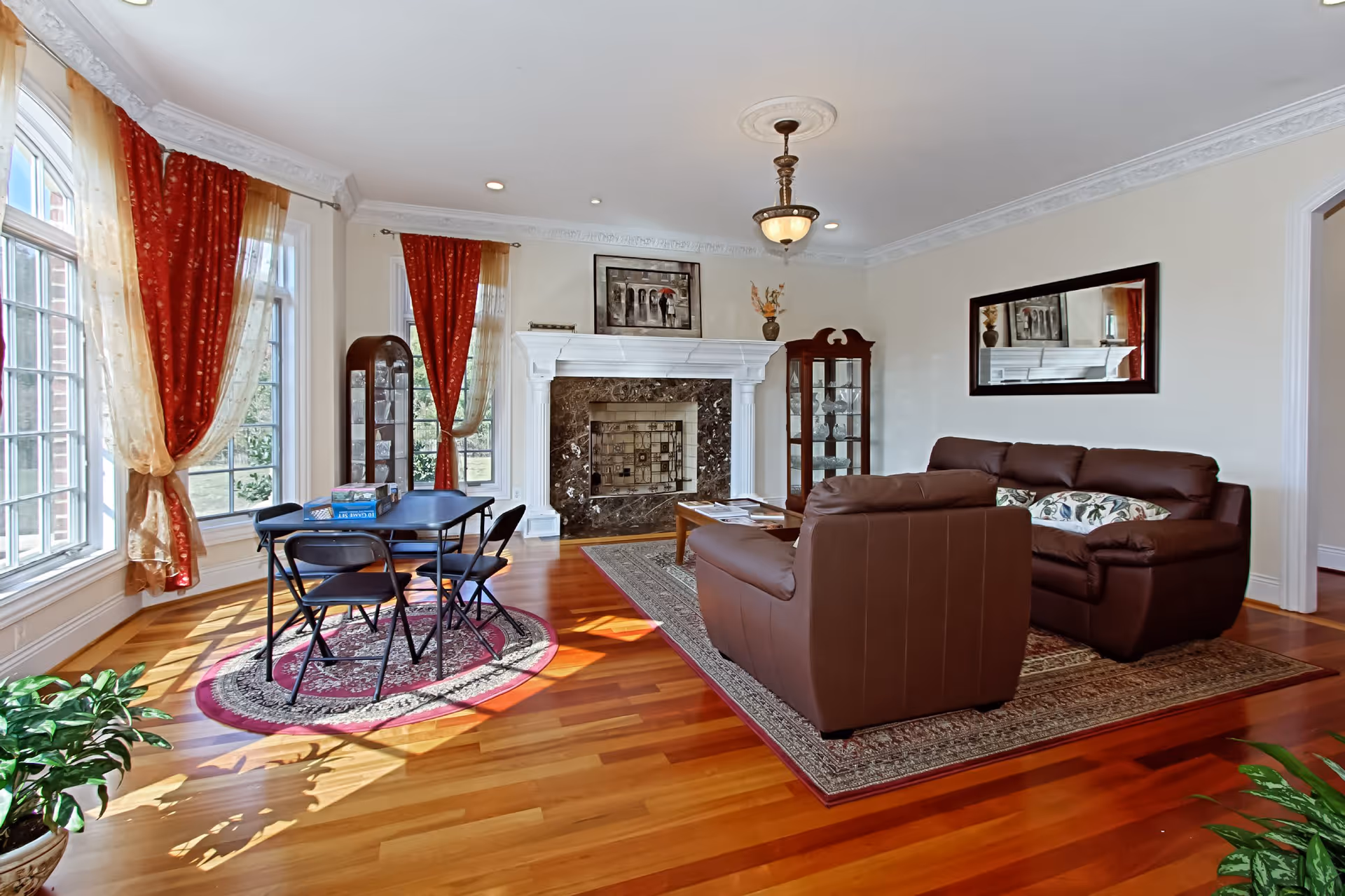 A bright living room with large windows covered by red and gold curtains, a dark brown leather sofa set on a patterned rug, a wooden coffee table, and a white fireplace with a dark marble surround. There are two glass display cabinets on either side of the fireplace, a large mirror on the wall, and a small table with four black folding chairs on a round rug near the windows. The room has hardwood floors and a ceiling light fixture.