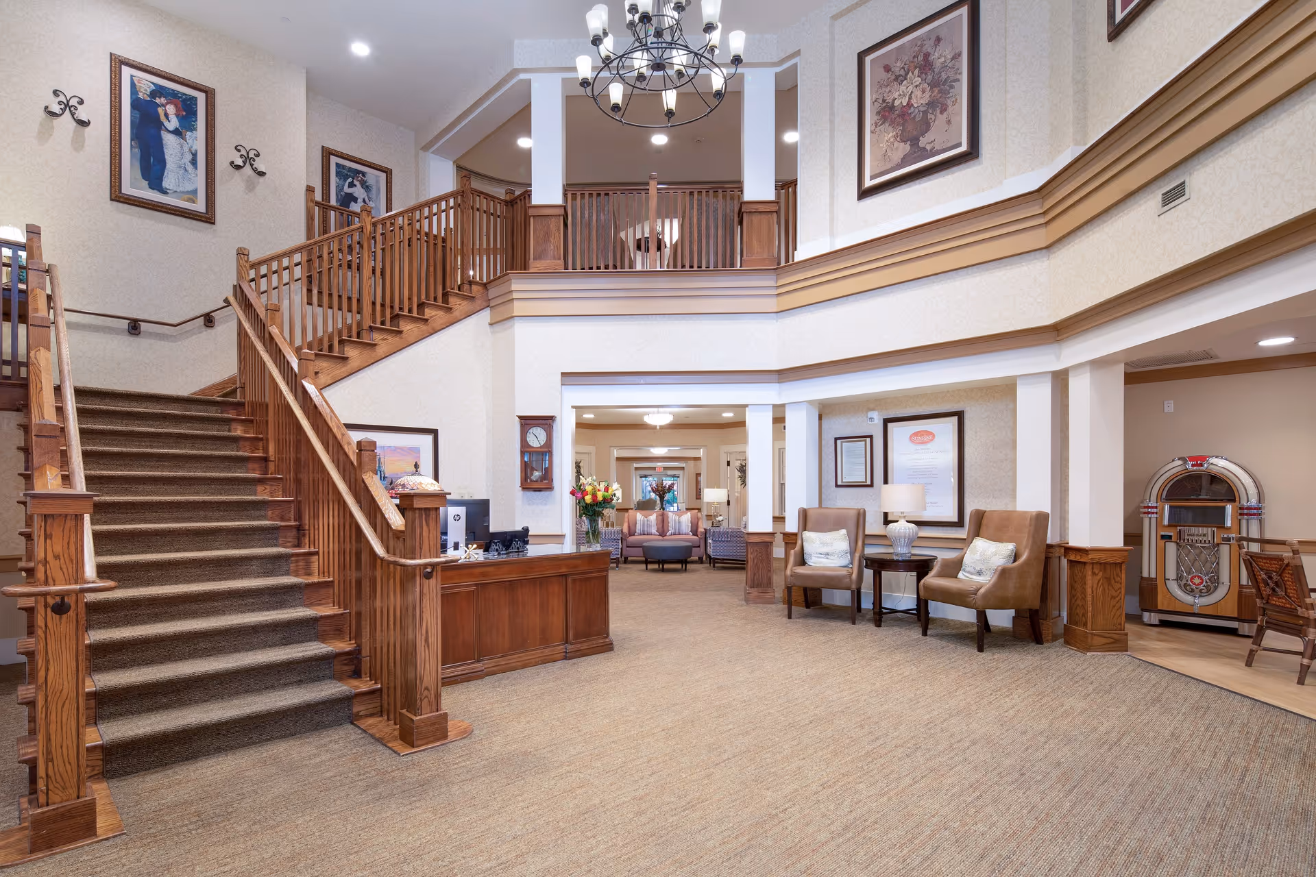 Spacious and well-lit interior of a senior living facility featuring a wooden staircase with carpeted steps, a wooden reception desk with a computer, comfortable seating area with two armchairs and a side table with a lamp, framed artwork on the walls, a vintage jukebox in the corner, and a chandelier hanging from the ceiling.