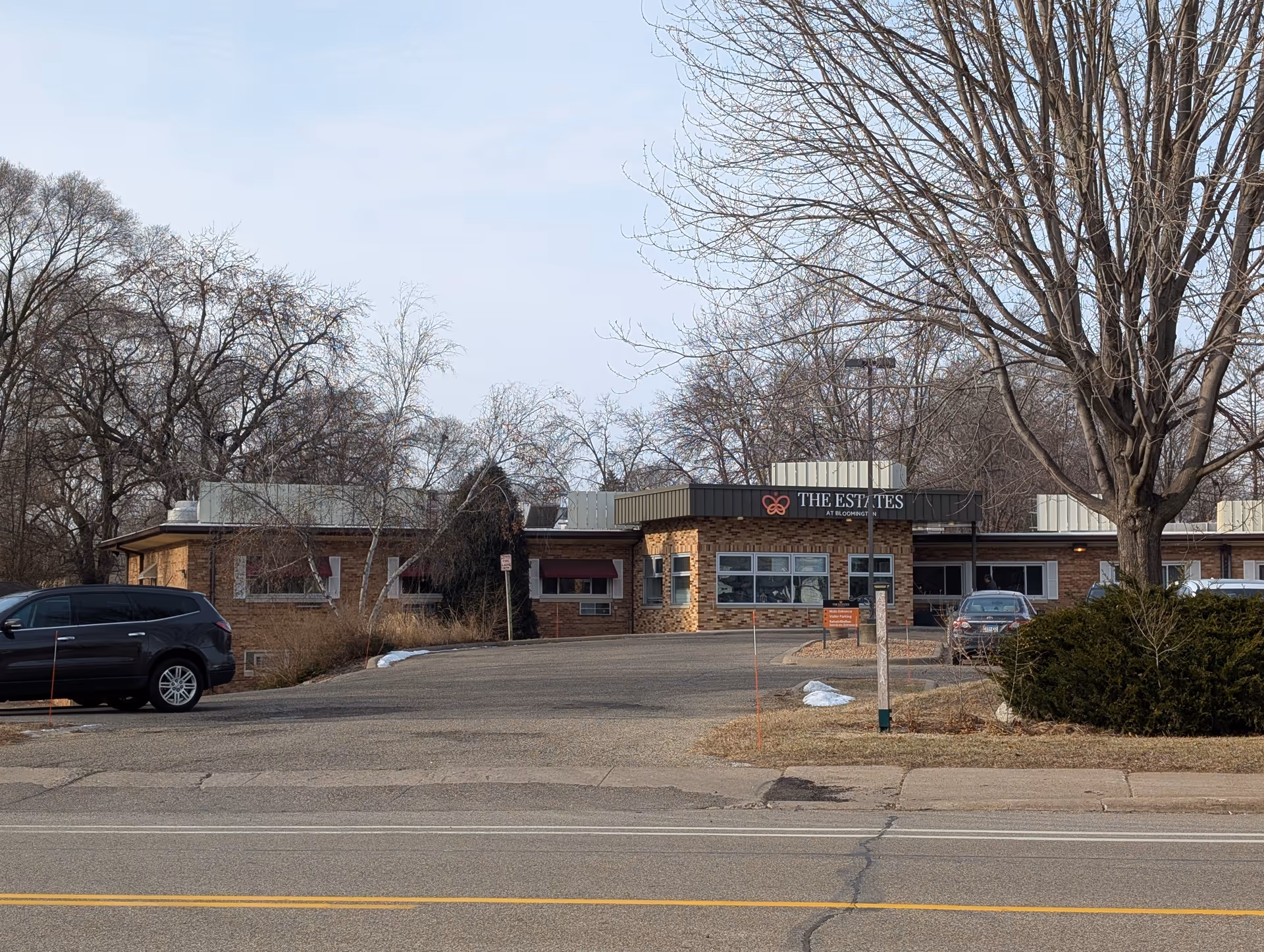 Front exterior view of The Estates at Bloomington building with a driveway, parked cars, leafless trees, and a clear sky.