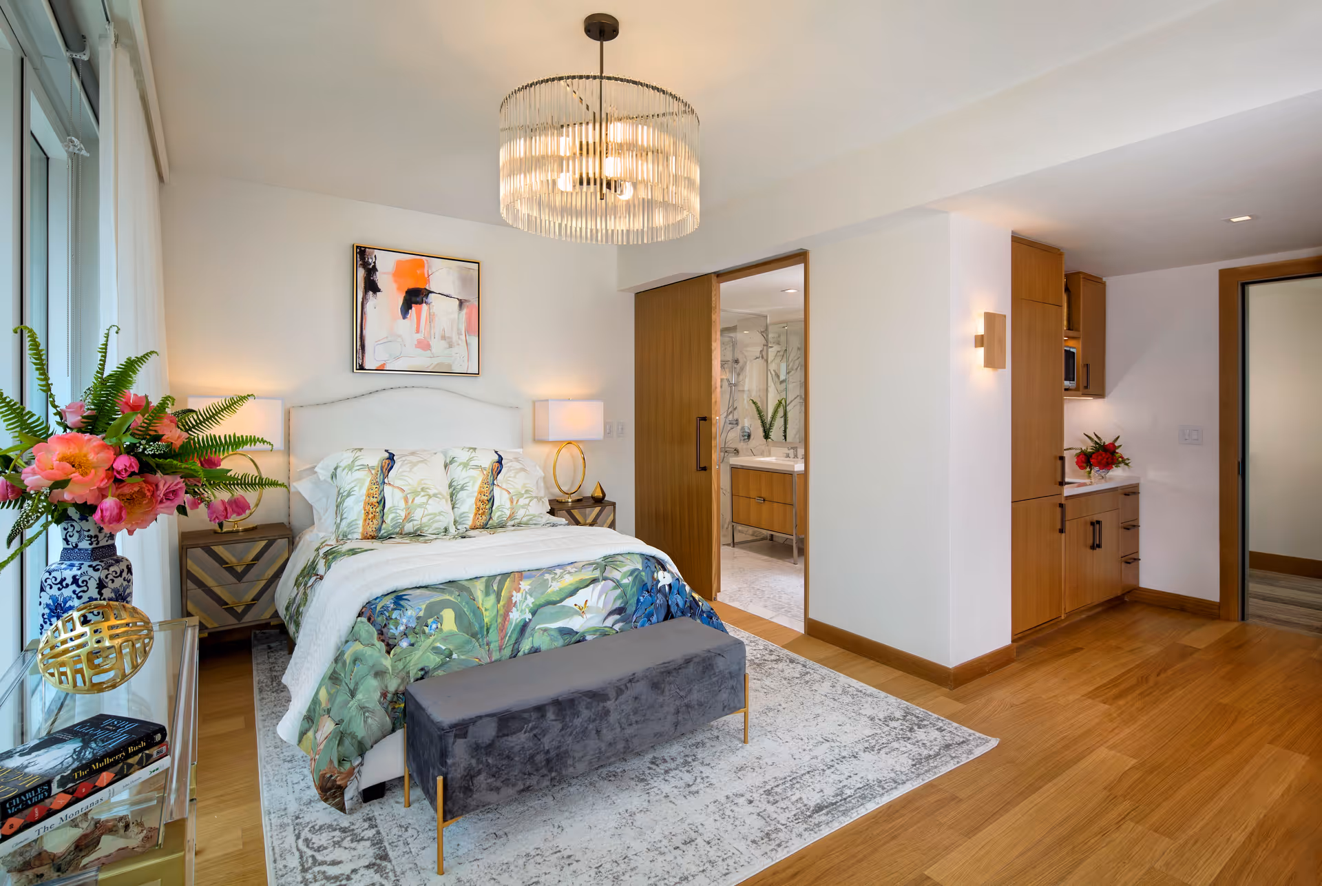 A bright and modern bedroom with a large bed featuring tropical-themed bedding and pillows. There is a gray bench at the foot of the bed, a patterned area rug, and a glass side table with a vase of colorful flowers and books. The room has wooden flooring and a contemporary chandelier hanging from the ceiling. To the right, there is an open doorway leading to a bathroom with a glass shower and wooden vanity. Adjacent to the bathroom is a small kitchenette area with wooden cabinets and a countertop with a flower arrangement.