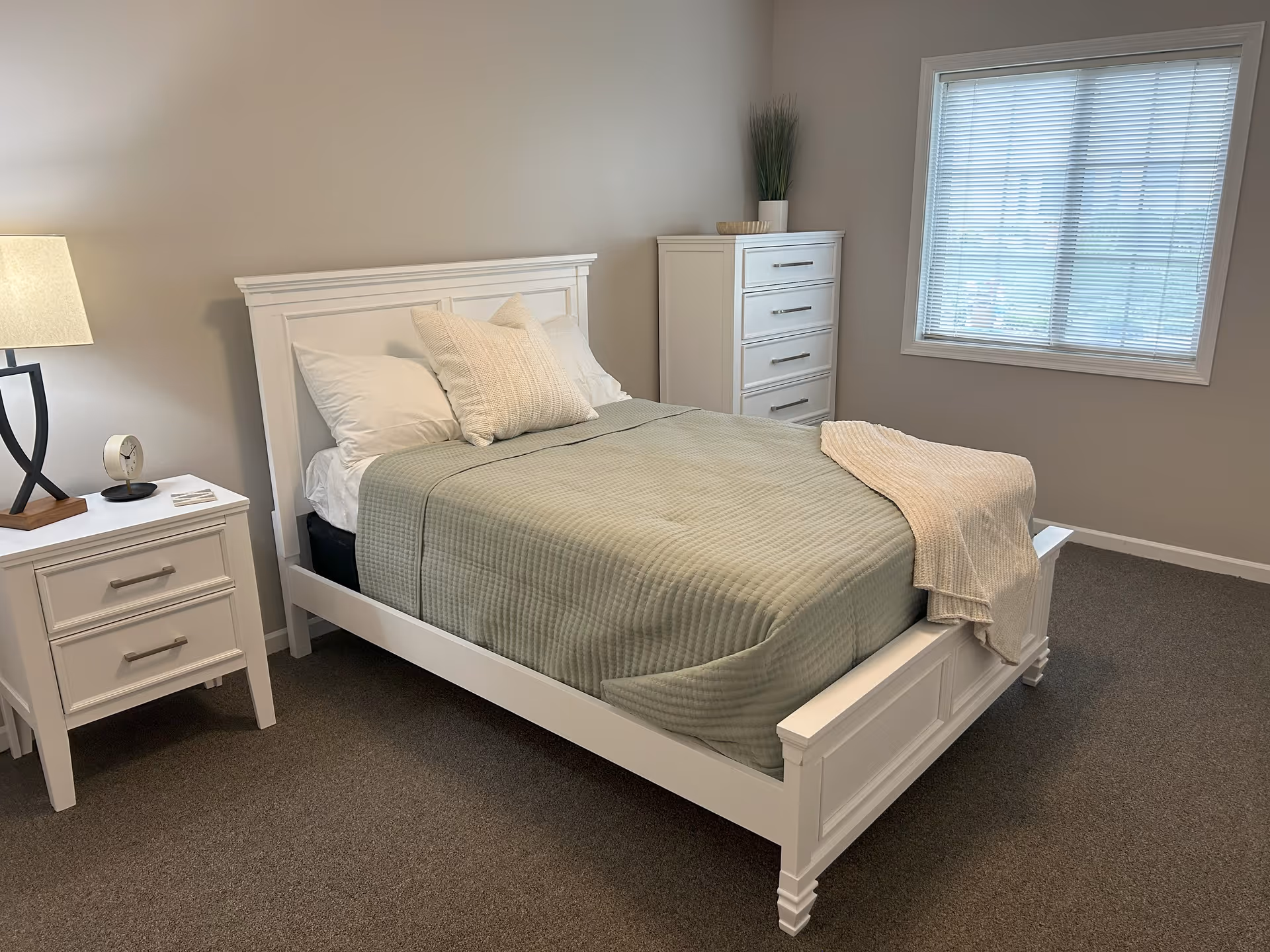 A neatly made bed with a white wooden frame, light green quilt, and white pillows in a bedroom. Next to the bed is a white nightstand with a lamp and a small clock. There is a white chest of drawers in the corner and a window with blinds letting in natural light.