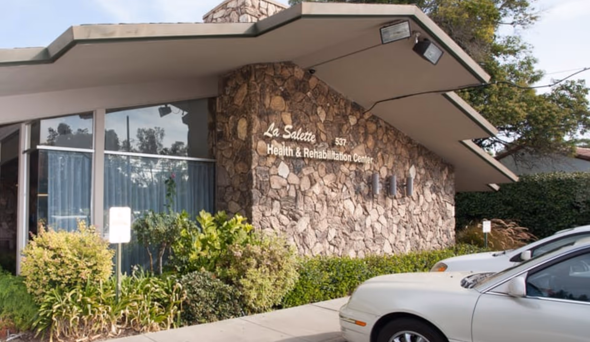 Exterior view of a health and rehabilitation center with a stone facade and large windows. The building has a sign that reads 'La Salette Health & Rehabilitation Center'. There are bushes and plants in front of the building and two cars parked nearby.