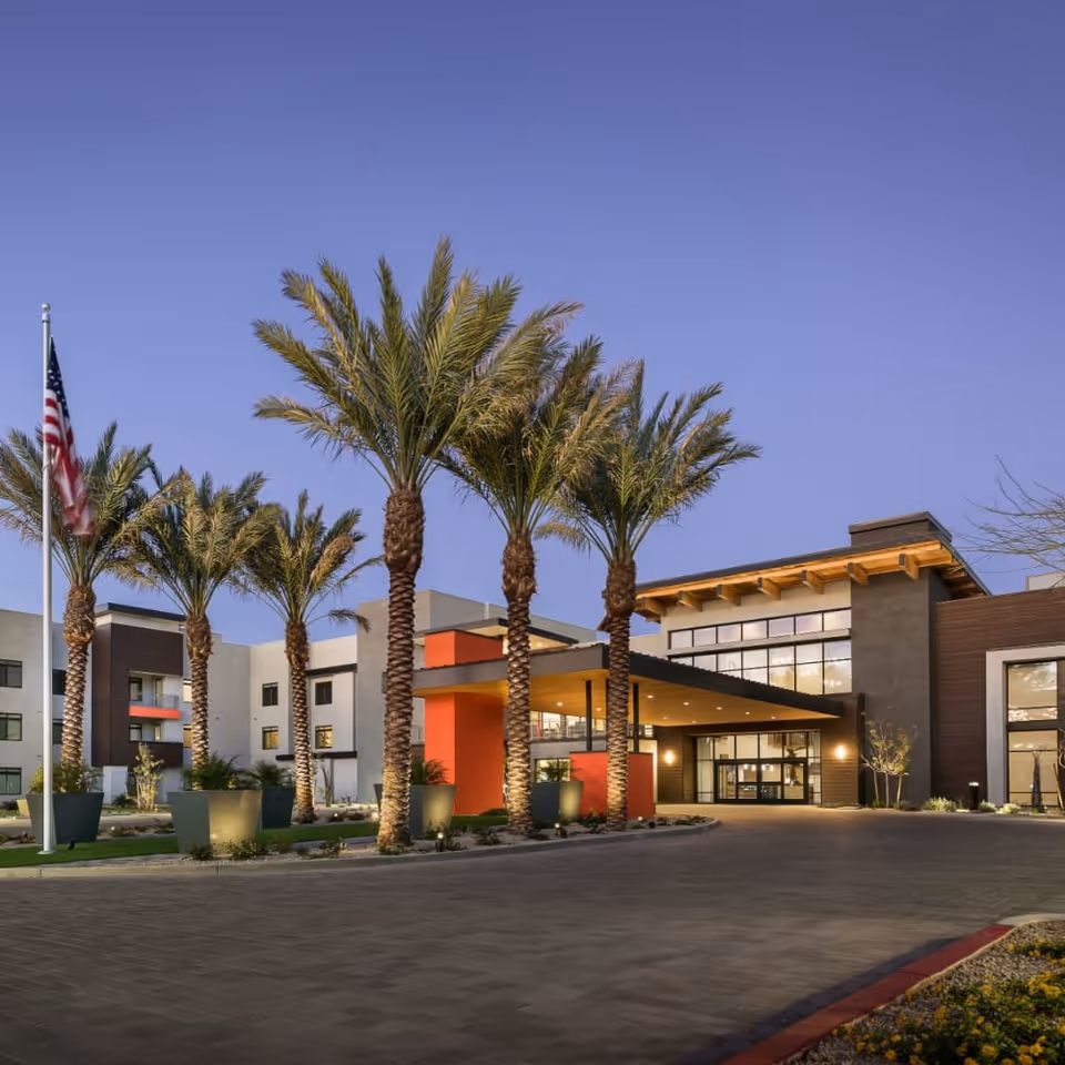 Front entrance of a modern senior living building at dusk with palm trees, an American flag, and an illuminated canopy.