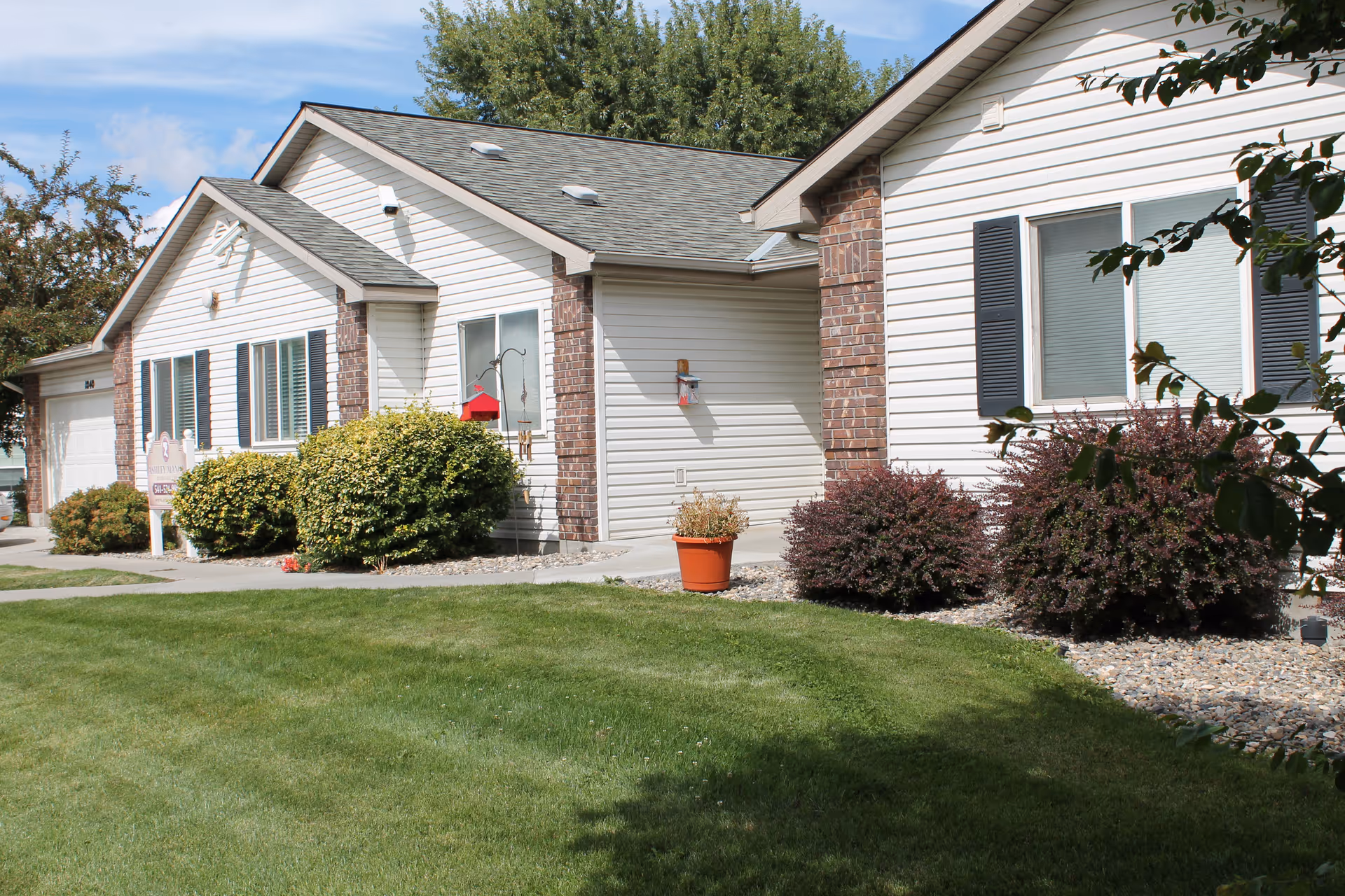 Exterior view of a single-story senior living facility building with white siding and brick accents, surrounded by green grass, bushes, and trees under a partly cloudy sky.