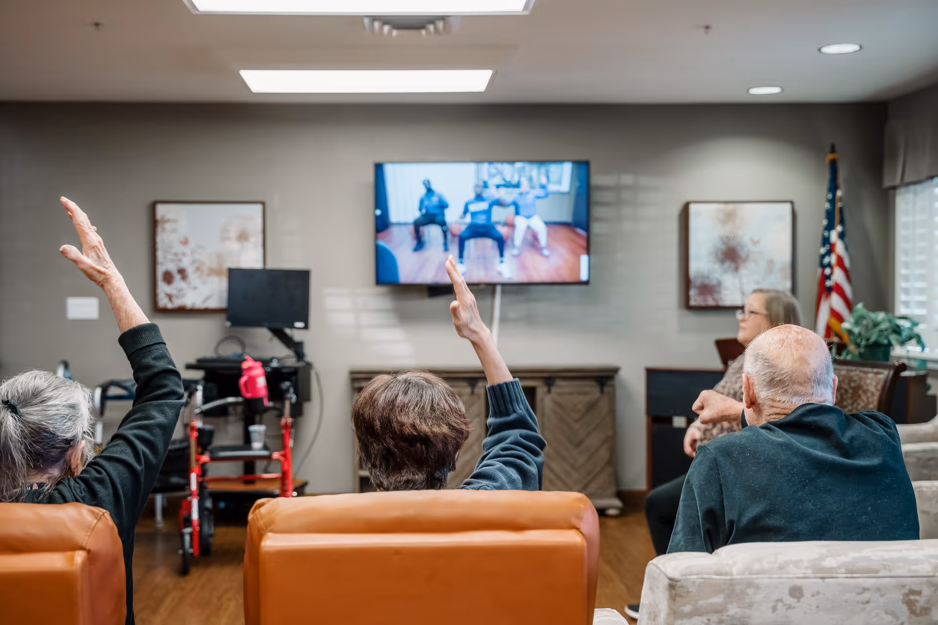 Several elderly residents seated in a common lounge raising their hands while following a fitness video on a wall-mounted TV.