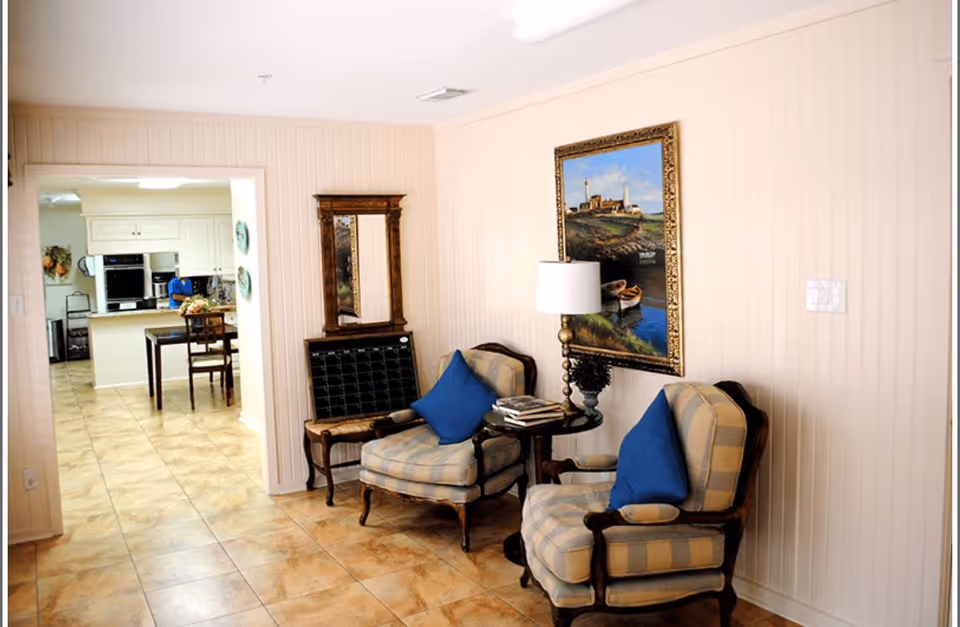 A cozy sitting area with two cushioned armchairs featuring blue pillows, a small round table with a lamp and books, a framed painting of a lighthouse on the wall, and a wooden mirror above a small bench. The area opens into a kitchen with a dining table and chairs, white cabinetry, and tiled flooring throughout.