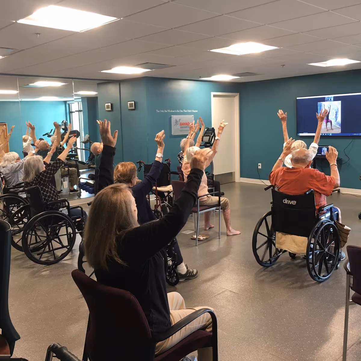 A group of elderly individuals, some in wheelchairs and others seated on chairs, participate in a seated exercise session in a room with teal walls and a large mirror. They are raising their arms while following an exercise video displayed on a wall-mounted TV screen.