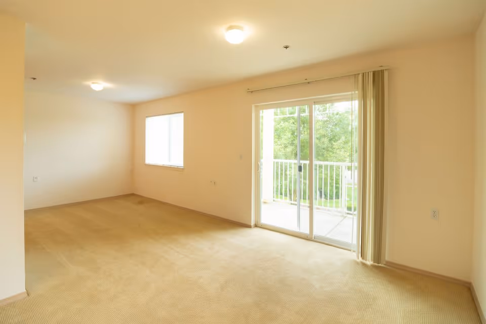 Empty room with beige carpet and cream-colored walls featuring a window and a sliding glass door leading to a balcony with a white railing and green trees outside.