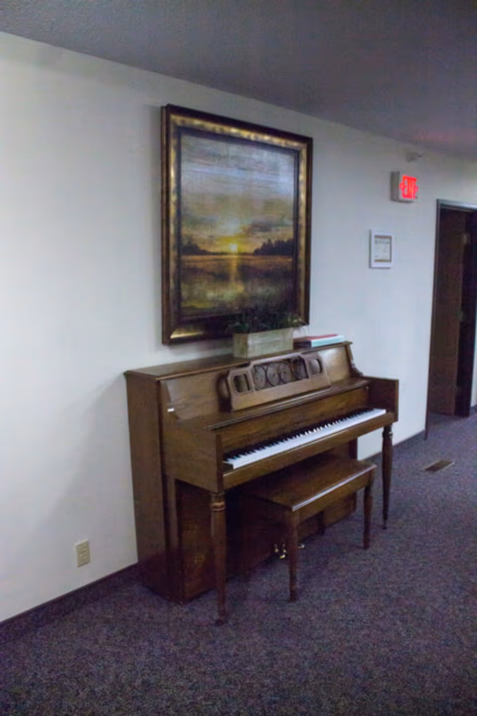 A wooden upright piano with a matching bench placed against a white wall in a carpeted hallway. Above the piano hangs a framed painting of a sunset over a body of water with trees in the background. There is a small plant and some books on top of the piano. An exit sign and a doorway are visible in the background.